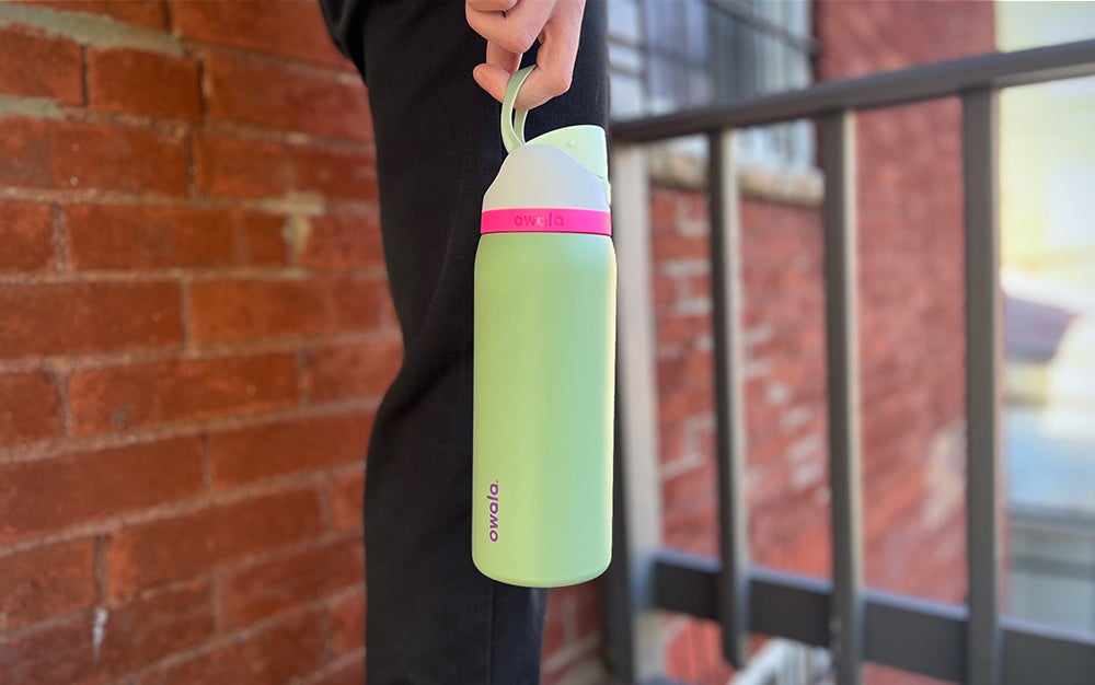 A person holding a green and pink Owala FreeSip water bottle in front of a brick wall.